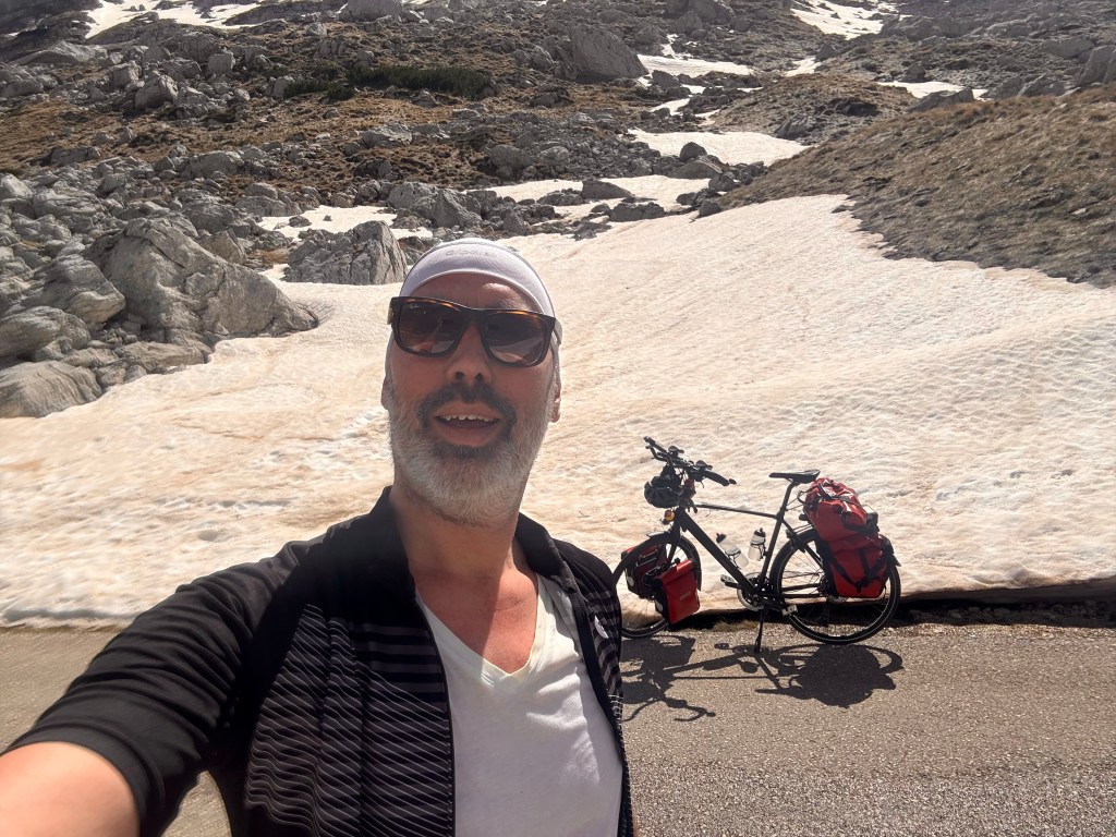 Daan Russchen over fietsen na kanker. Een man maakt een selfie met een fiets op een bergweg, omringd door sneeuw en rotsen.