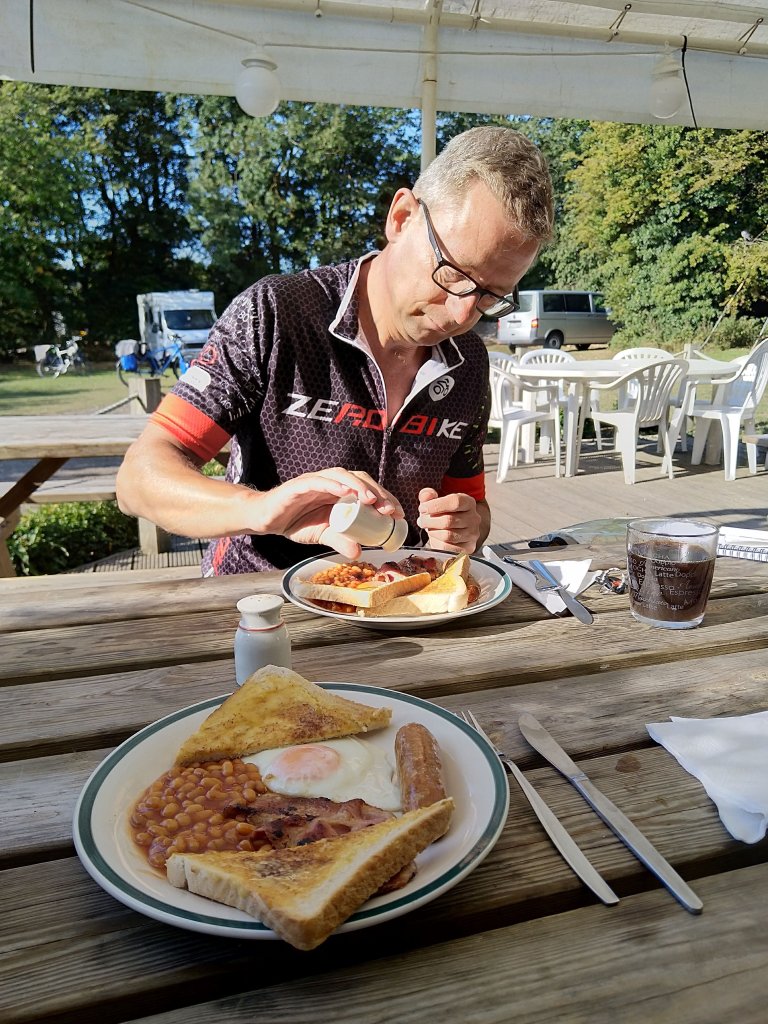 Brood voor fietsvakantie. Een man eet ontbijt aan een houten tafel, met een bord vol voedsel, waaronder eieren, worst, bonen en toast. Hij strooit kruiden over zijn eten terwijl er een kopje koffie naast hem staat.