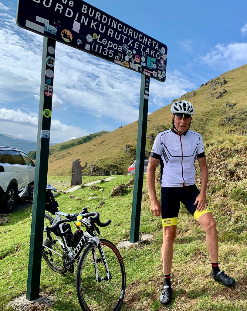 een man met wit wielershirt met fiets op een berg bij bord Burdincurucheta (Lepoa) 1135m