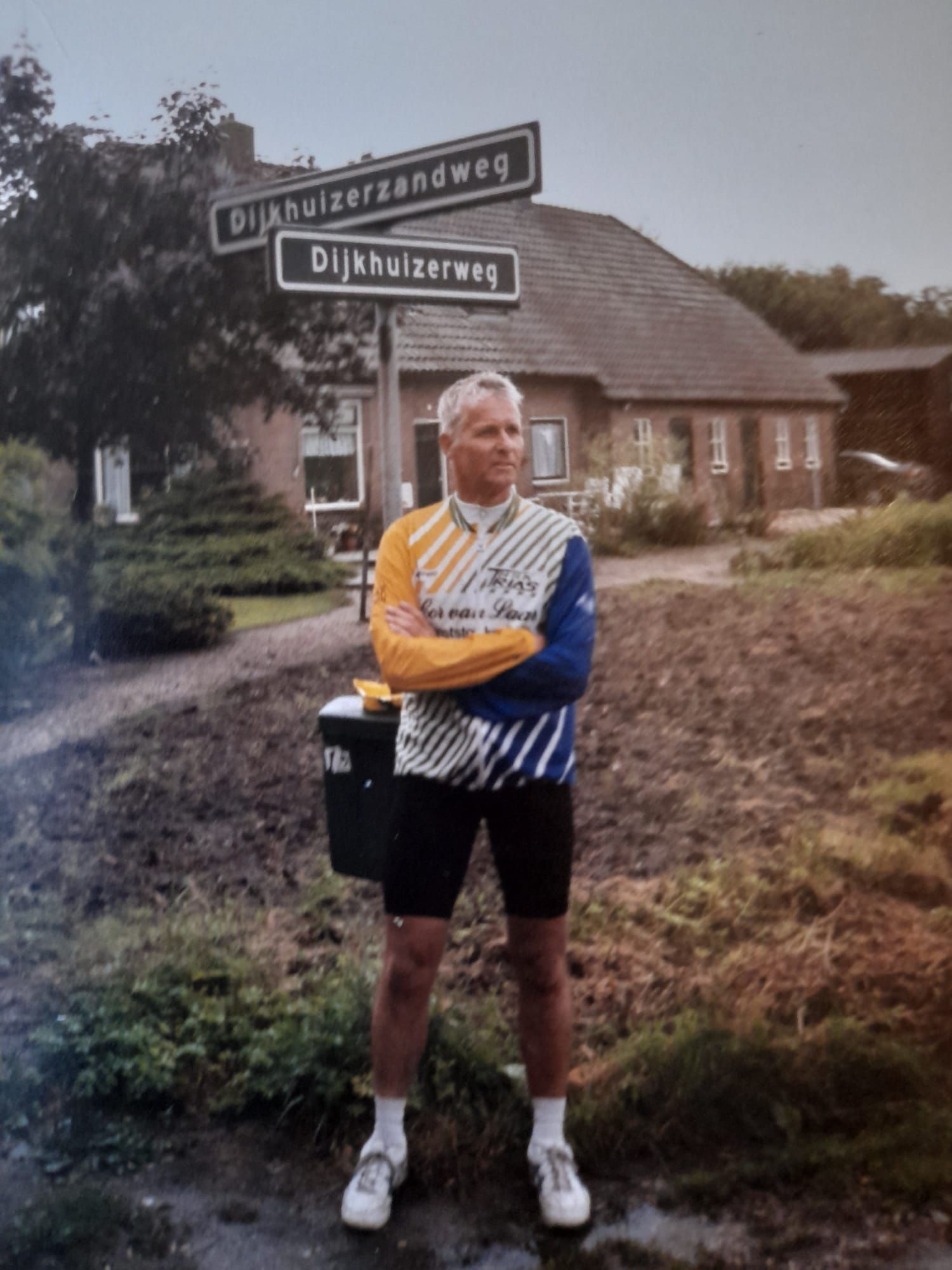 Martin Dijkhuizen: vrijheid, eenvoud en rust op twee wielen. Man wearing colorful cycling jersey and shorts standing with arms crossed near street signs reading Dijkhuizerzandweg and Dijkhuizerweg in a rural setting