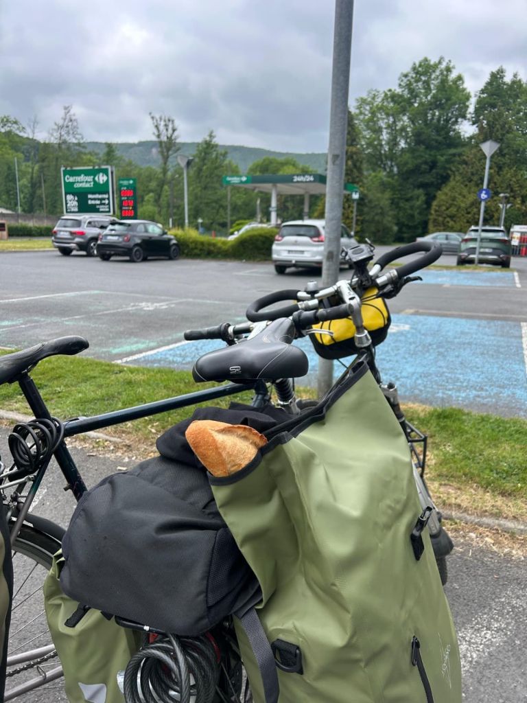 Thuis op de fiets. Een fiets met een groene fietstas waarin een stokbrood zichtbaar is, geparkeerd op een parkeerplaats naast een tankstation met een groene omgeving en bewolkte lucht.