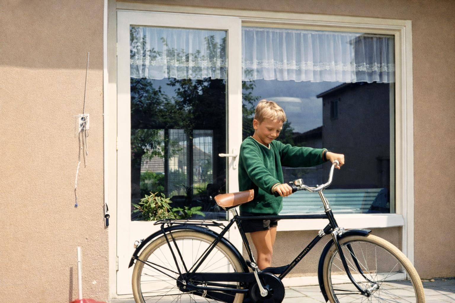 A young boy in a green sweater standing outside a house, holding the handlebars of a black bicycle with beige tires